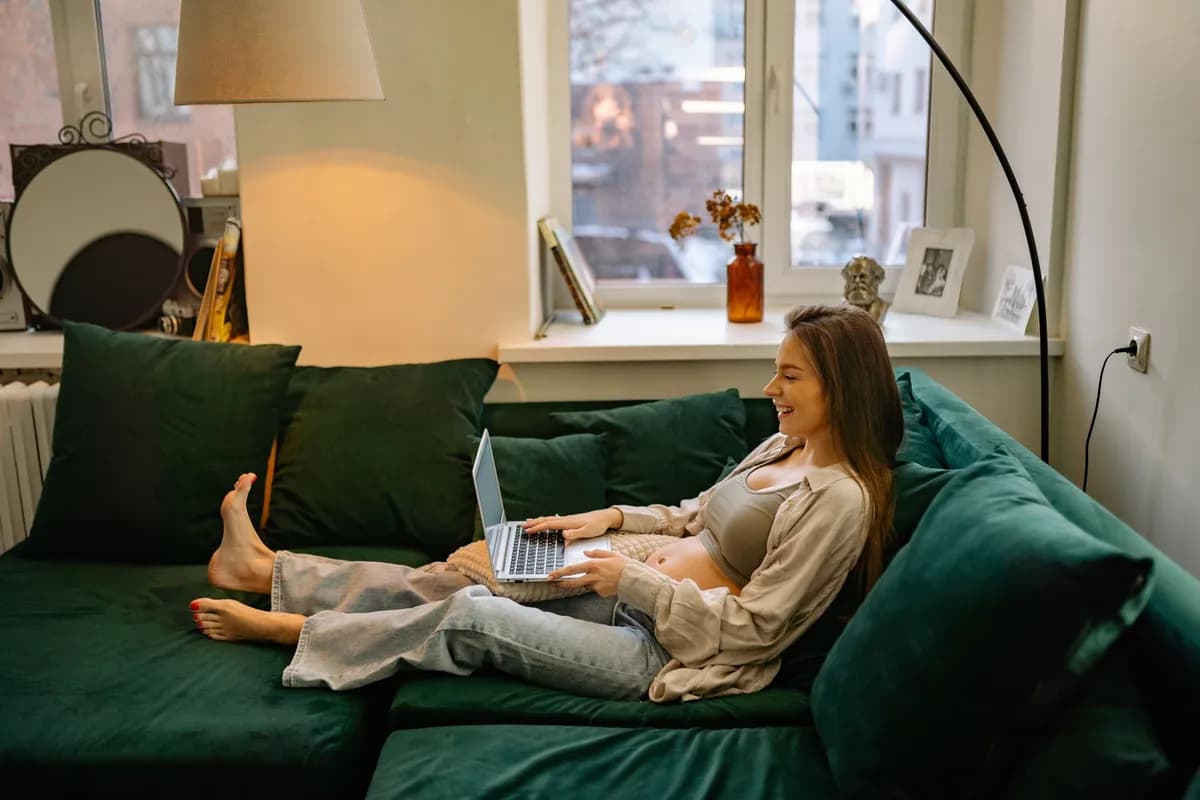 Zwangere vrouw op de bank met een laptop op schoot bij het raam